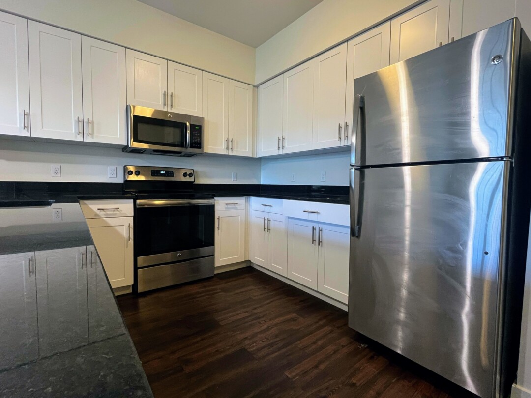 A kitchen with white cabinets and dark countertops, stainless steel appliances including a refrigerator, oven, and over-the-range microwave, and dark wood flooring that adds warmth and contrast.