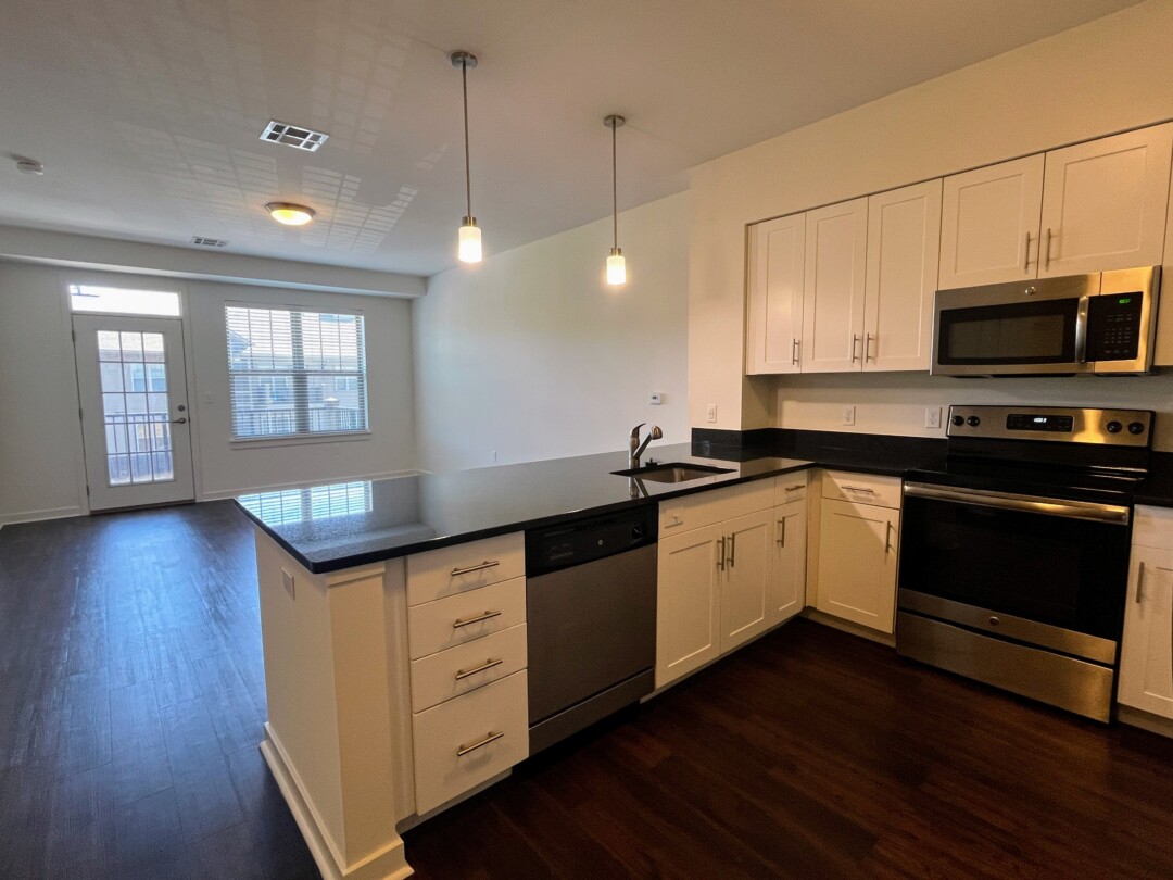 A kitchen with white cabinets and dark countertops, stainless steel appliances including a refrigerator, oven, dishwasher, and over-the-range microwave, a sink set into a counter with two hanging light fixtures above, and dark wood flooring that extends into an open living area with a window and glass door.