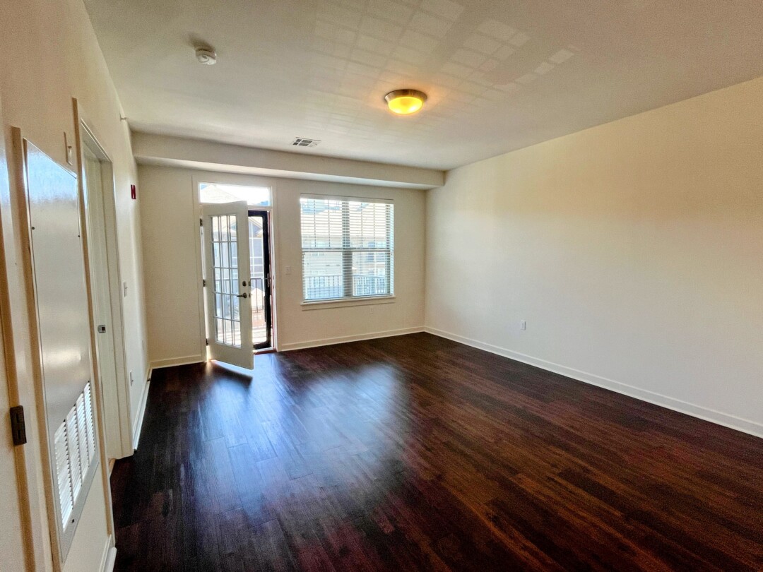 An empty living area with dark wood flooring and white walls, featuring a window with blinds and a glass door leading outside that is partially open, with ceiling-mounted lighting and a doorway visible along the side wall.