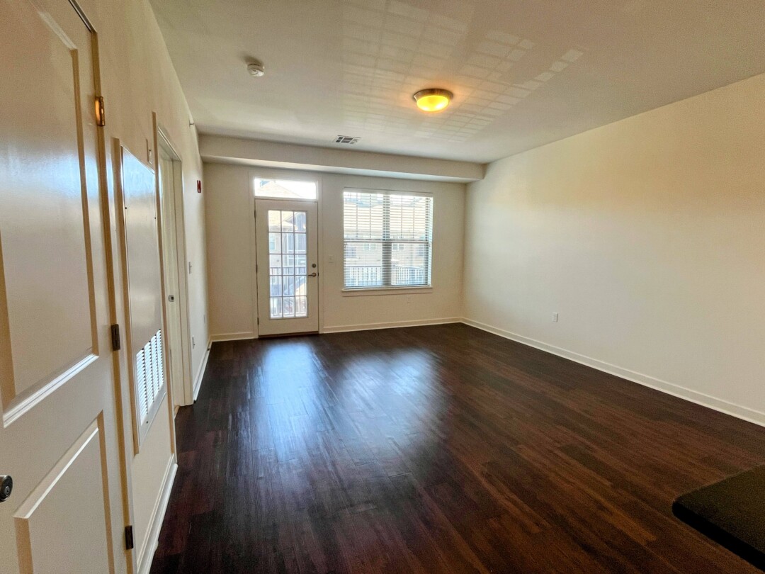 An empty living area with dark wood flooring and white walls, featuring a window with blinds and a glass door leading outside, with ceiling-mounted lighting and a doorway visible along the side wall.