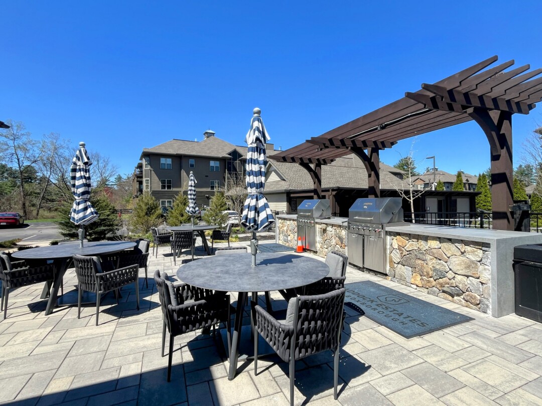 An outdoor patio area with round tables and chairs, each shaded by striped umbrellas, set on a paved surface, with built-in stainless steel grills along a stone counter, a wooden pergola overhead, and residential buildings and trees in the background under a clear blue sky.