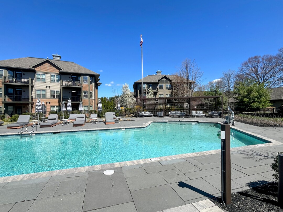 A swimming pool with clear blue water surrounded by lounge chairs and umbrellas on a paved deck, with a fenced seating area, landscaped surroundings, and residential buildings in the background under a clear blue sky.