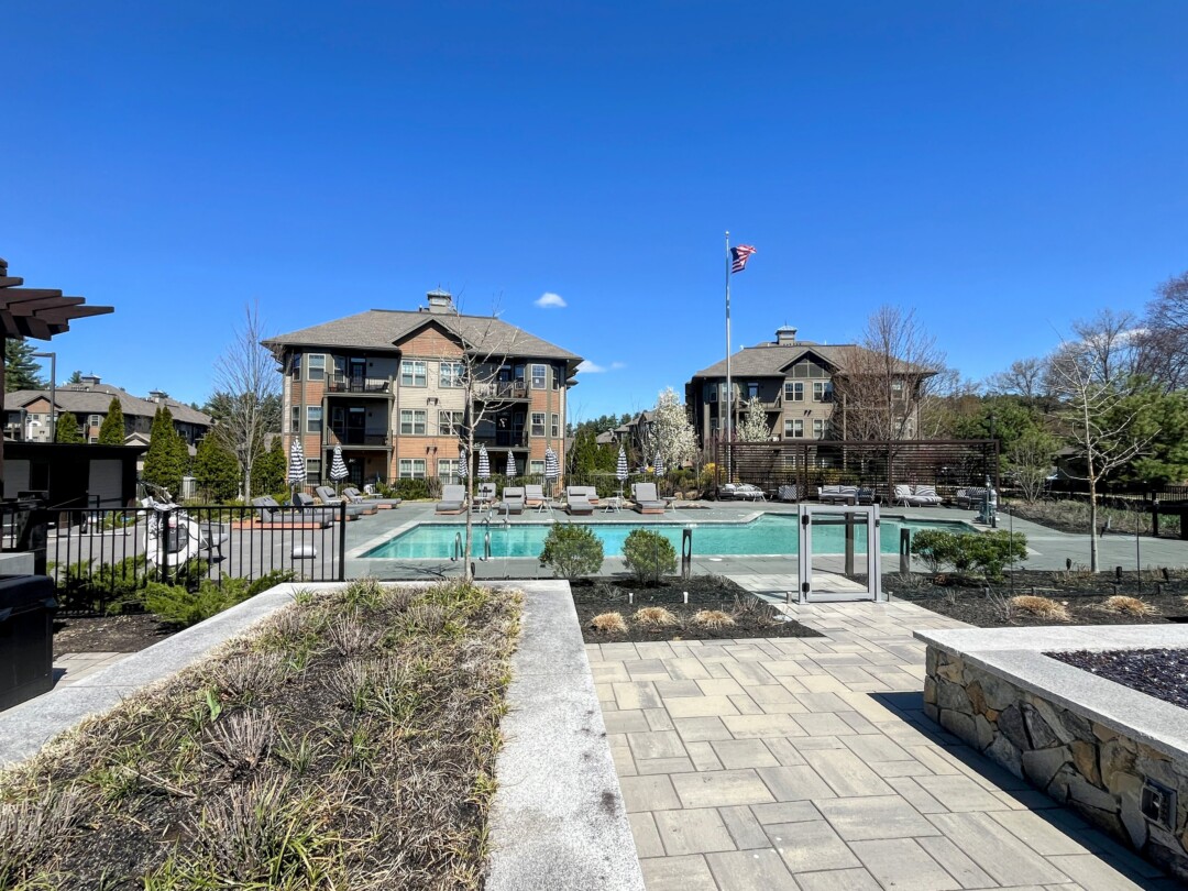 An outdoor courtyard with a swimming pool surrounded by lounge chairs and umbrellas, landscaped with plants and paved walkways, with residential buildings in the background and an American flag on a flagpole under a clear blue sky.