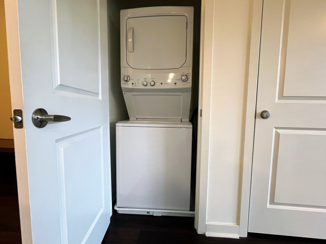 A stacked washer and dryer unit inside a small closet with white doors, set against a light-colored wall with dark wood flooring.