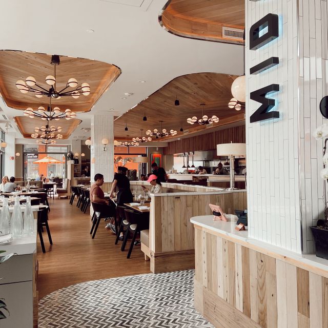 Restaurant interior with wood flooring, tables and chairs, booth seating, wood-paneled accents, white ceiling with wood inset sections, and hanging light fixtures.