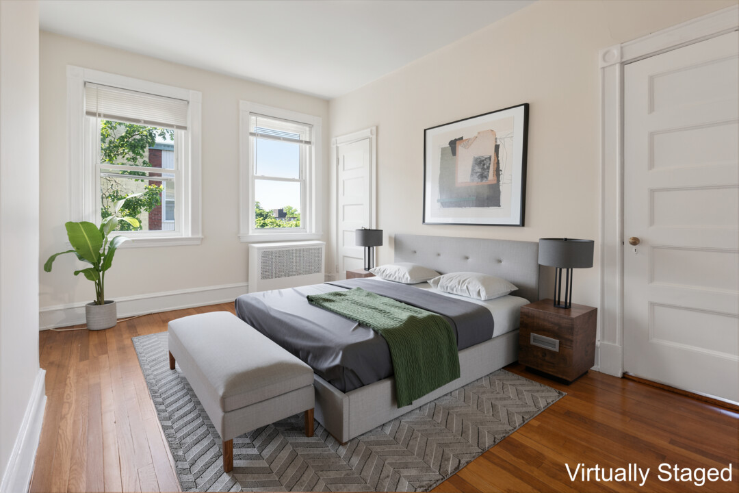 Bedroom with white walls, hardwood flooring, two windows, a white door, and furniture including a bed, nightstands, bench, rug, and table lamps.