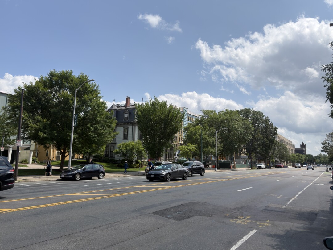 Wide paved roadway with multiple traffic lanes, yellow center markings, and rows of mature green trees. Low-rise residential buildings and cars line the street beneath a bright blue sky with scattered white clouds.