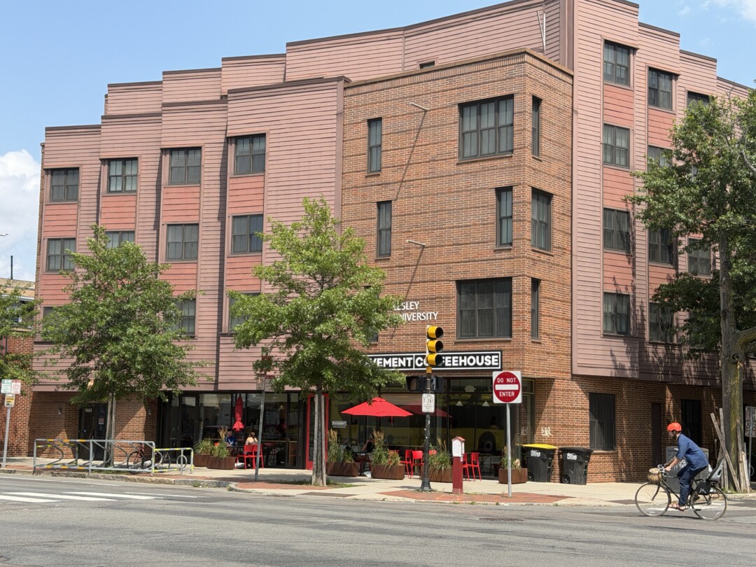 Modern four-story building with red and brown brick exterior, large windows, and a ground-floor storefront with outdoor seating.