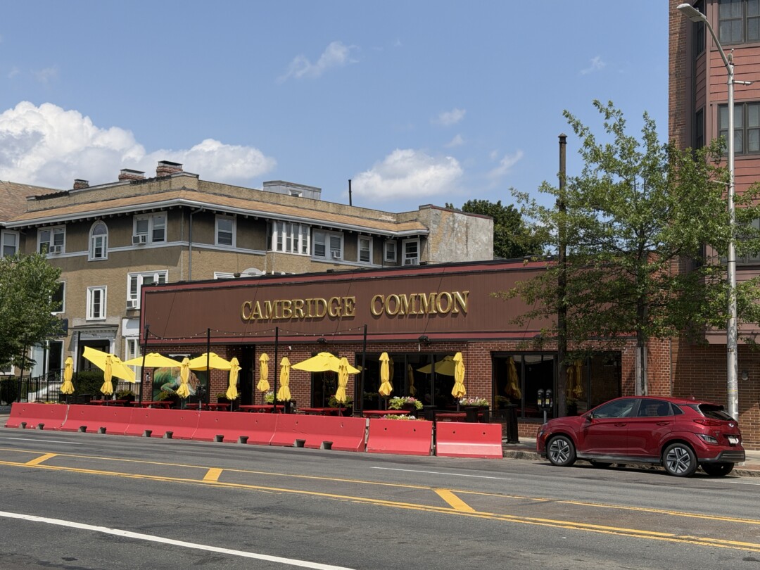 Single-story brick restaurant with dark red awnings, bright yellow patio umbrellas, and a red outdoor barrier along the sidewalk.