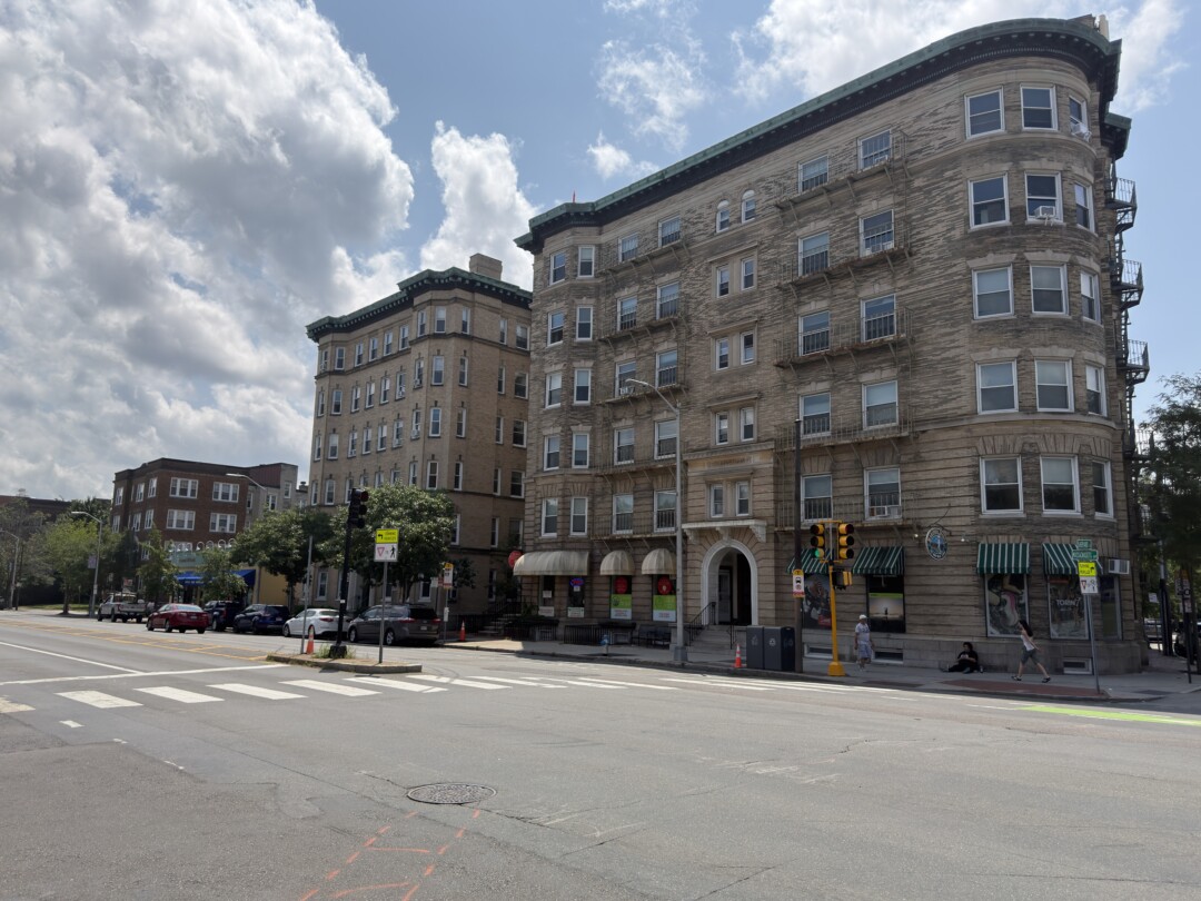 Light brown brick apartment building with rows of windows and ground-floor storefronts along a wide street under a partly cloudy sky.