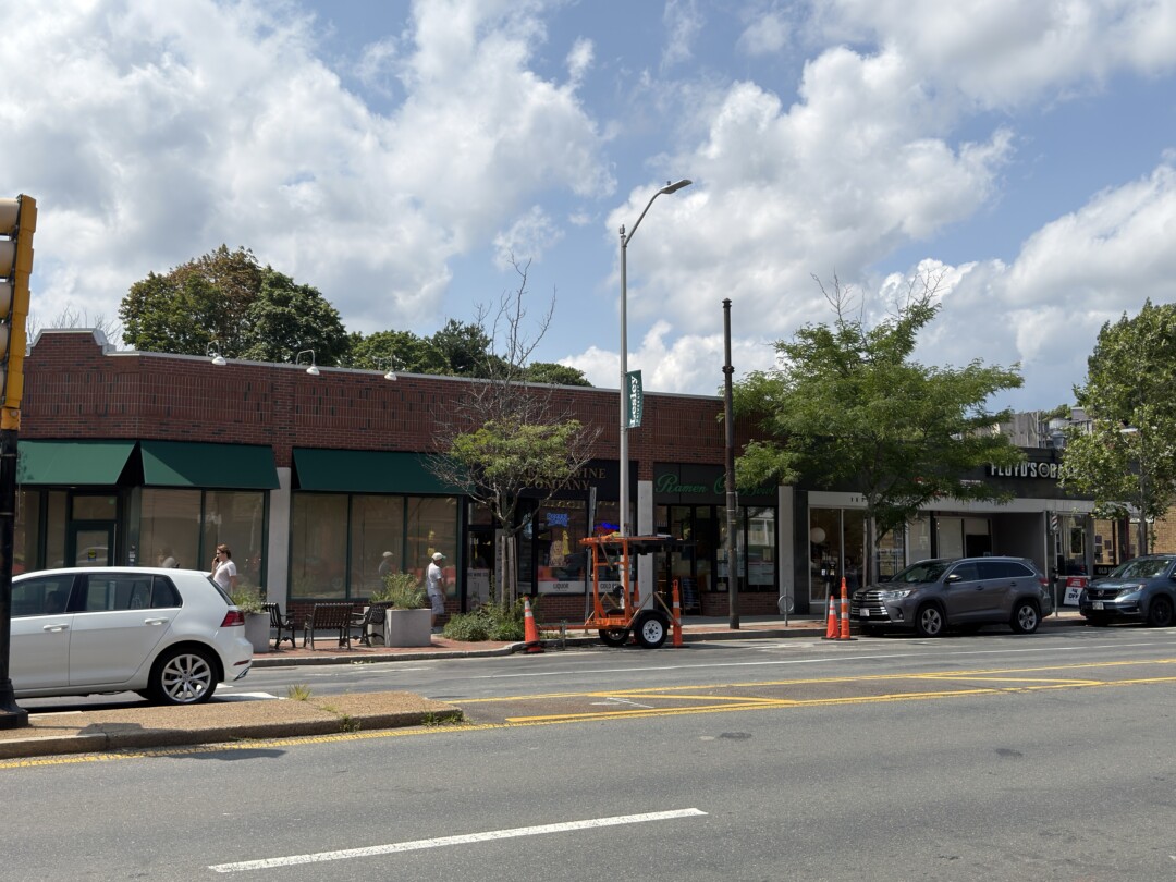 Row of one-story storefronts with brown brick and green awnings, large front windows, and a sidewalk facing a wide street.