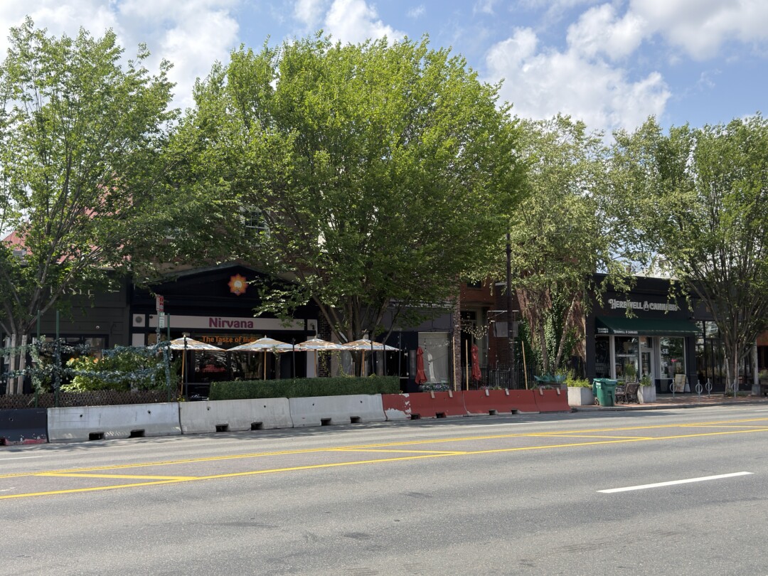 Commercial storefronts partially shaded by large green trees, with dark-colored façades, outdoor seating, and wide sidewalks.Commercial storefronts partially shaded by large green trees, with dark-colored façades, outdoor seating, and wide sidewalks.