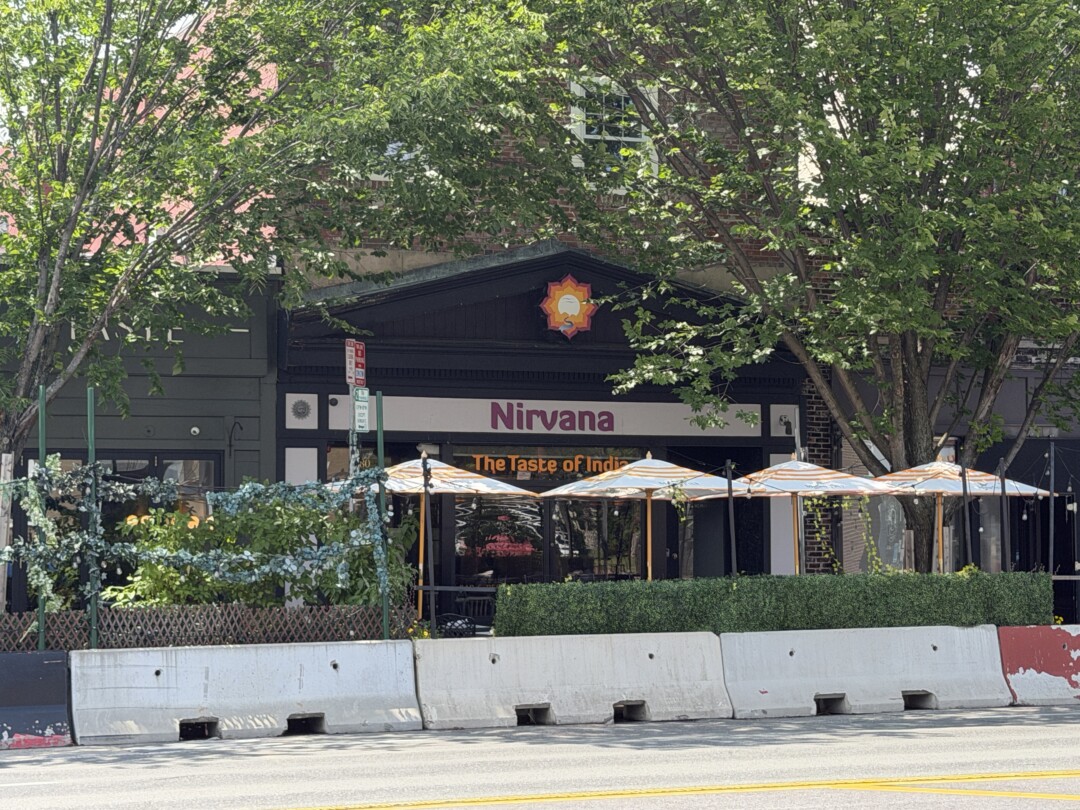 Commercial storefronts partially shaded by large green trees, with dark-colored façades, outdoor seating, and wide sidewalks.Commercial storefronts partially shaded by large green trees, with dark-colored façades, outdoor seating, and wide sidewalks.