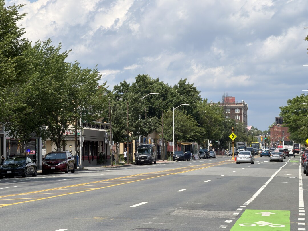 Broad multi-lane street with white traffic markings, a green painted bike lane, leafy trees, parked cars, and brick buildings extending into the distance under a cloudy blue sky.