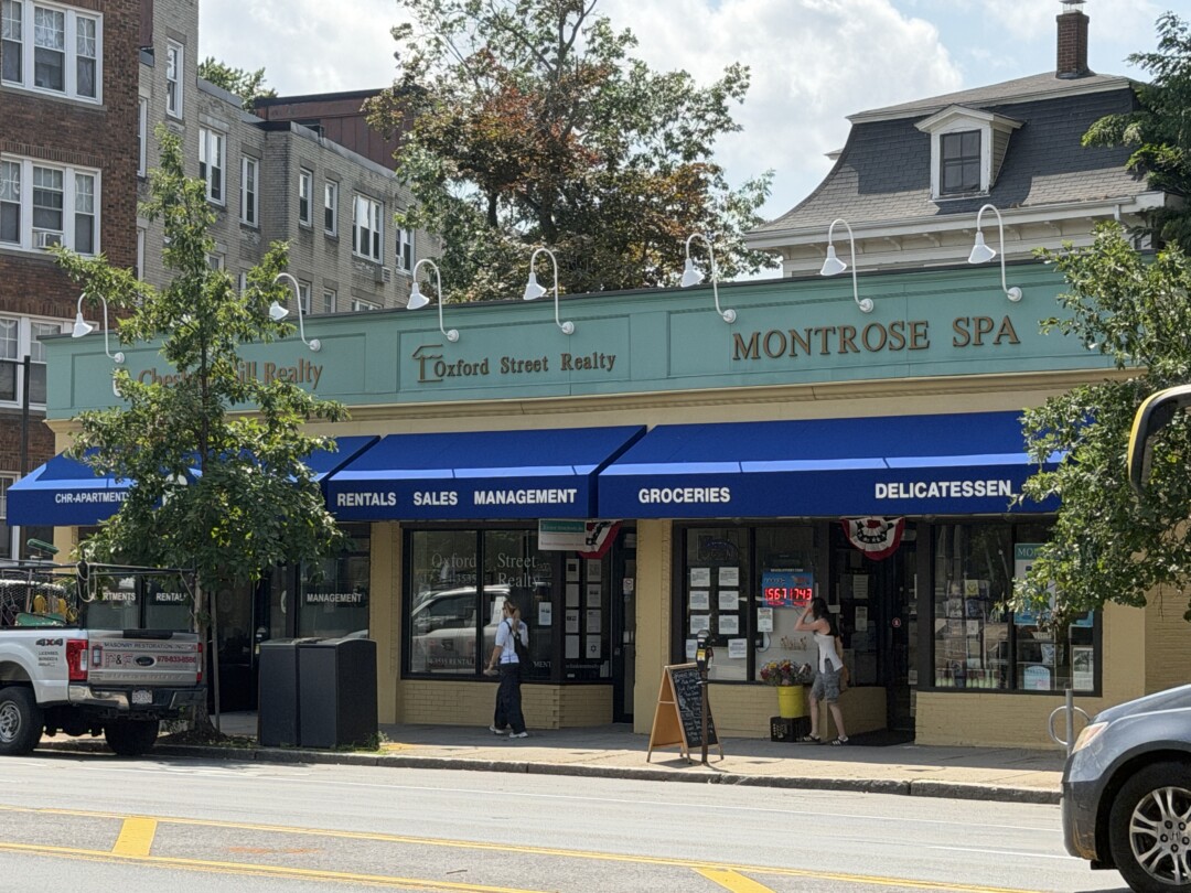 Closer view of beige commercial storefronts with blue awnings, glass windows, and adjacent brick buildings framed by leafy trees.
