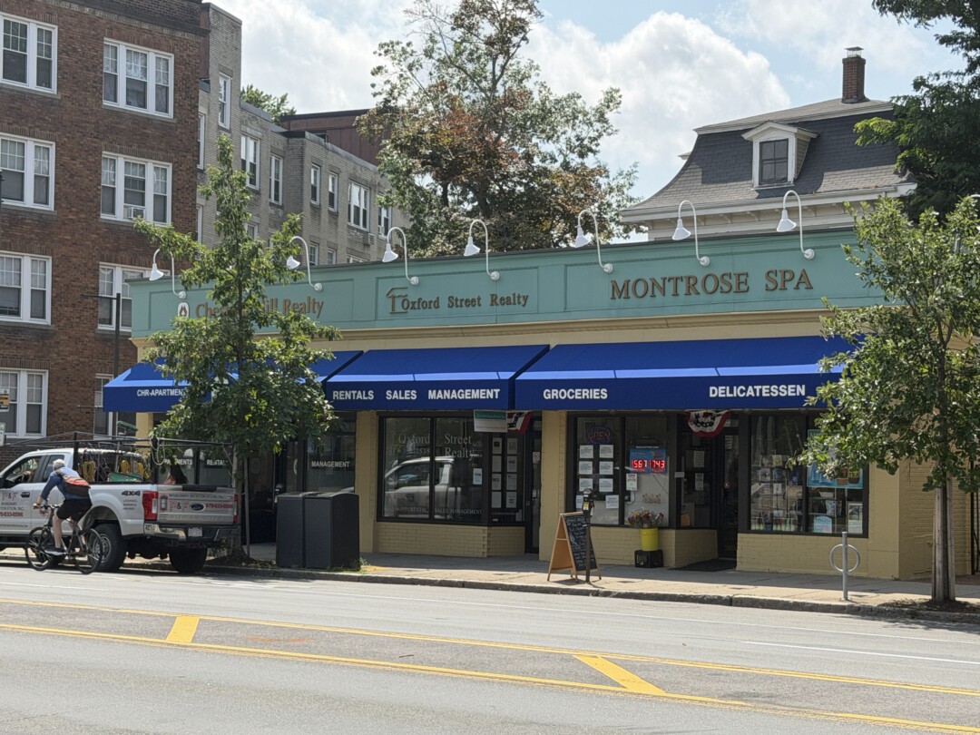 Commercial establishment with beige siding, blue awnings, large storefront windows, and neighboring brick residential buildings.