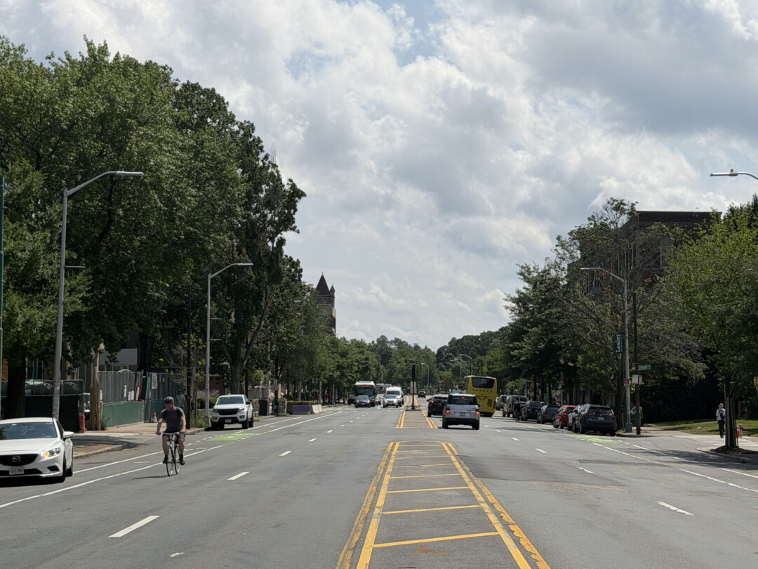 Wide multi-lane street with yellow road markings, rows of mature trees, parked vehicles, and a bright, partly cloudy sky.