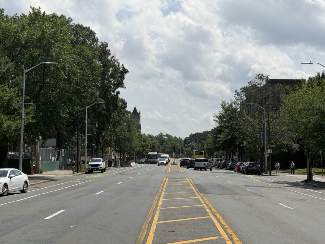 Wide multi-lane street with yellow road markings, rows of mature trees, parked vehicles, and a bright, partly cloudy sky.