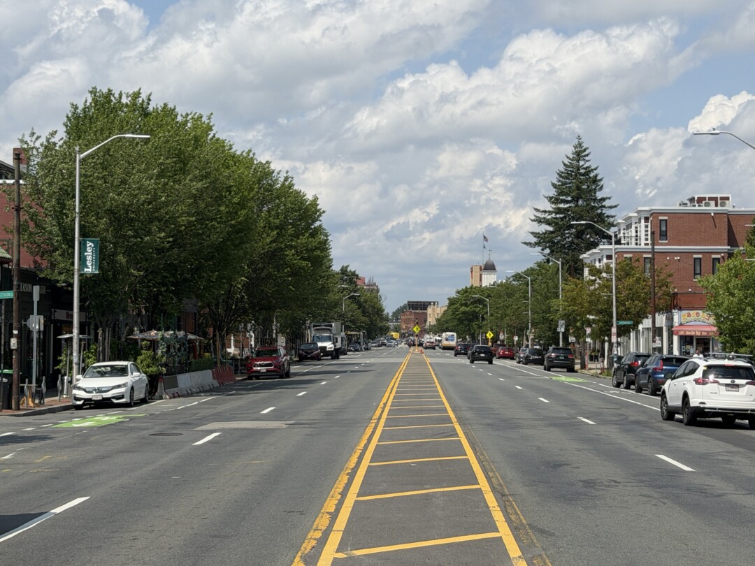 Wide multi-lane street with yellow road markings, rows of mature trees, parked vehicles, and a bright, partly cloudy sky.