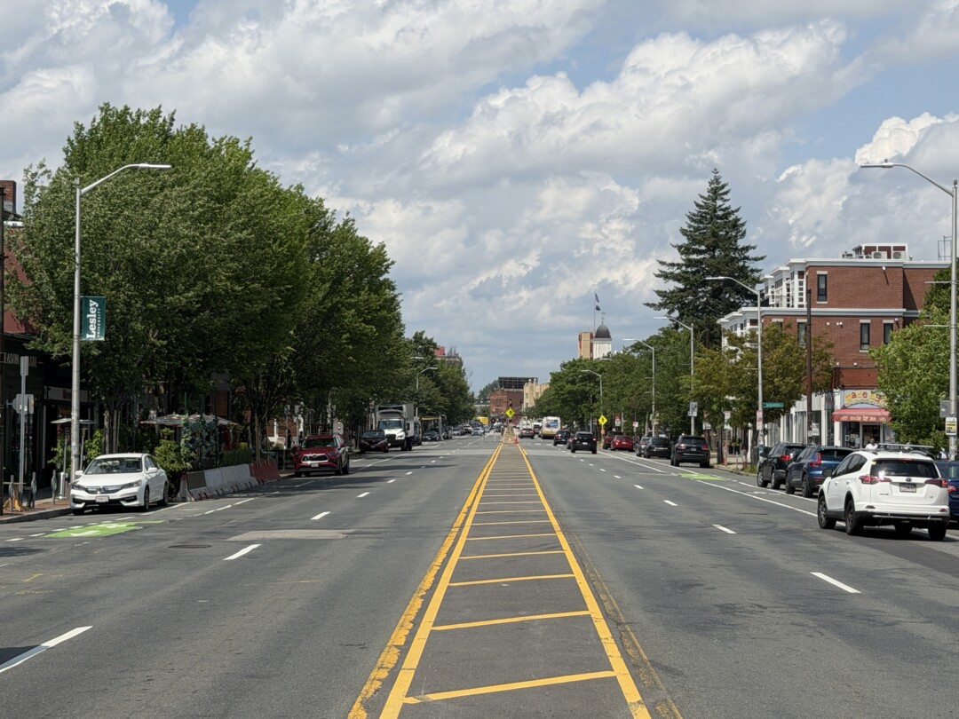 Wide multi-lane street with yellow road markings, rows of mature trees, parked vehicles, and a bright, partly cloudy sky.