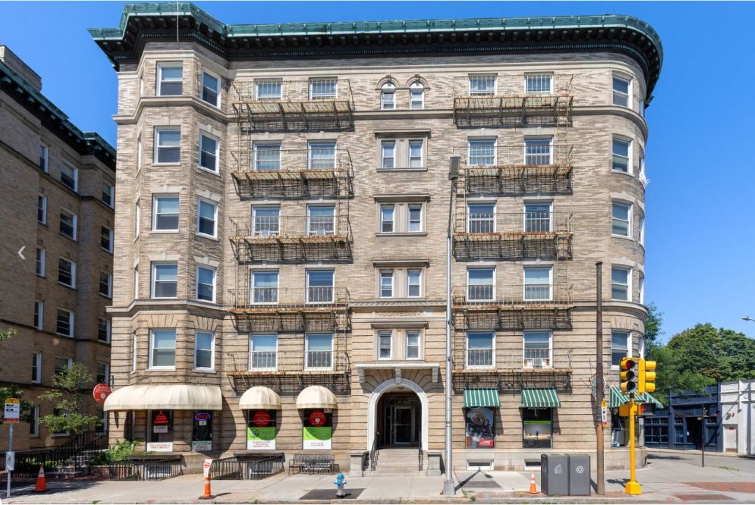 Exterior view of a multi-story brick apartment building with balconies, arched entrance, storefronts at street level, and large windows.