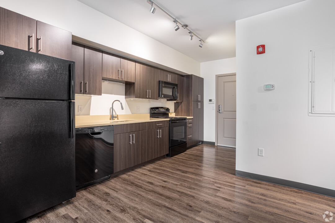 Open kitchen with dark cabinets, black appliances, wood-style flooring, white walls, and a high white ceiling.
