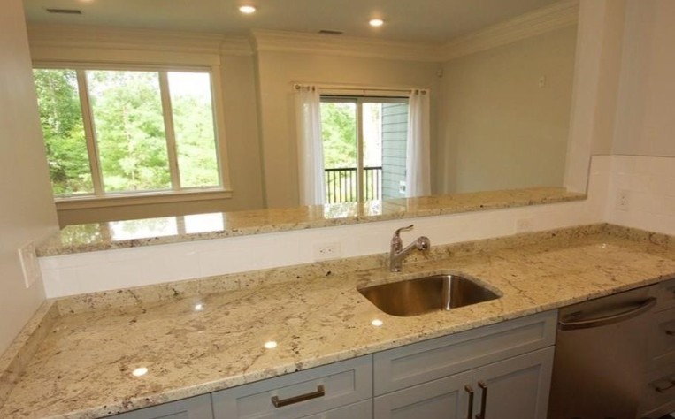 Kitchen with light stone countertops, a stainless steel sink, gray cabinets, stainless steel appliances, and views into the living room with large windows and a glass door.