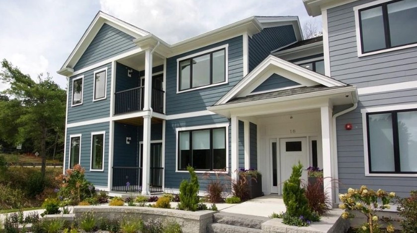 Two-story residential building with blue and gray siding, white trim, black railings, landscaped walkways, and large windows.