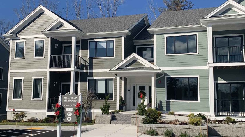 Two-story residential building with gray and green siding, white trim, black railings, large windows, landscaped grounds, and a community sign near the entrance.