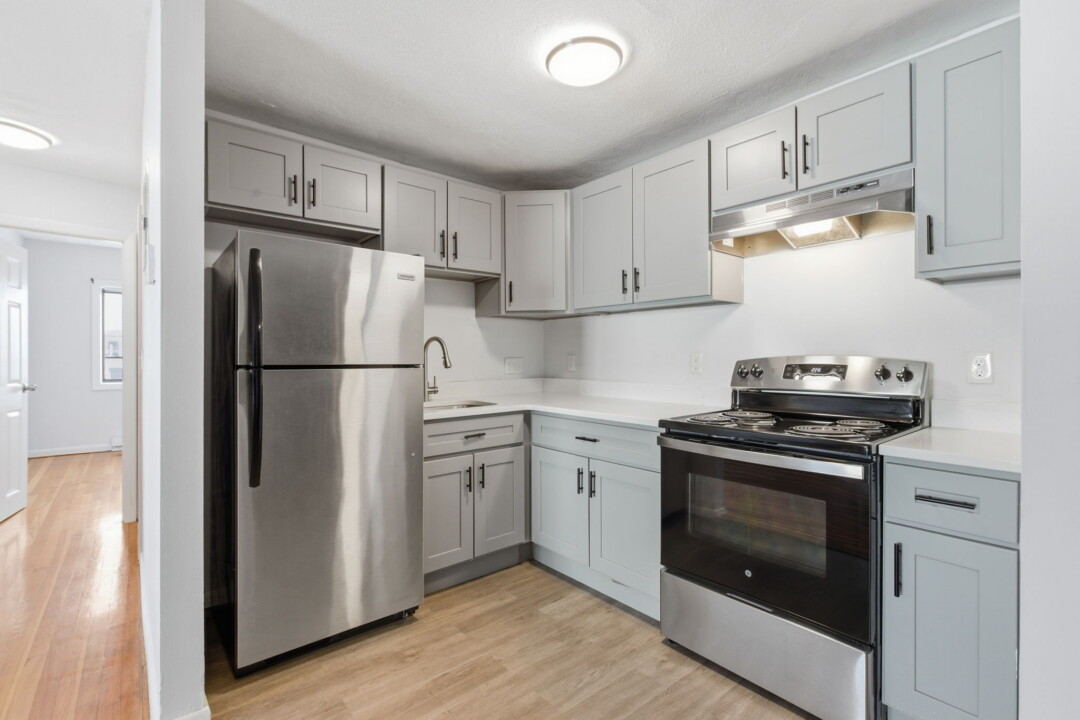 Compact kitchen with light gray cabinets, stainless steel refrigerator and stove, white countertops, and light wood flooring.