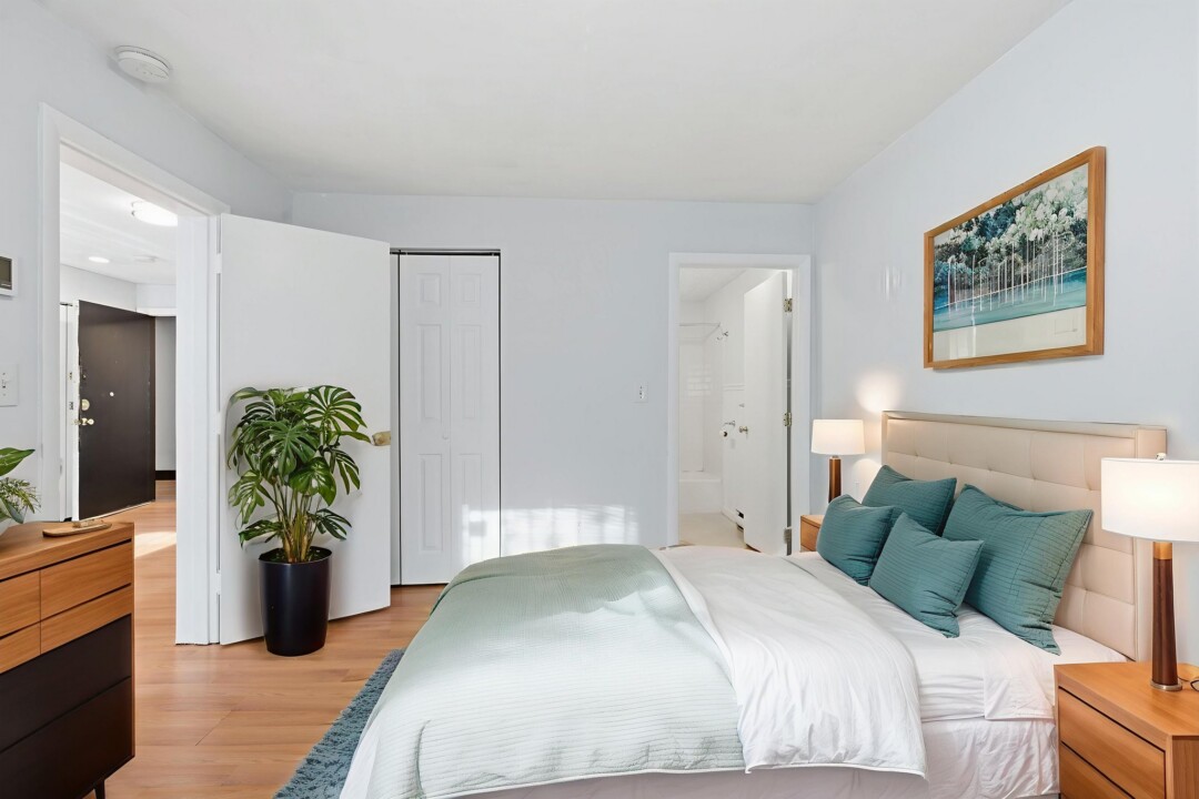 Bright furnished bedroom with a bed, bedside lamp, wood flooring, and a potted plant.