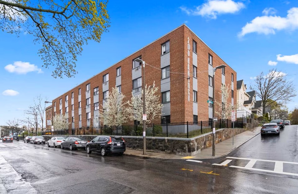 Exterior view of a multi-story brick residential building with large windows, trees, and street parking.