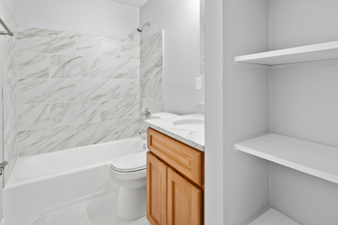 Bathroom with a bathtub and shower, toilet, wood vanity, marble-patterned walls, and built-in shelving.