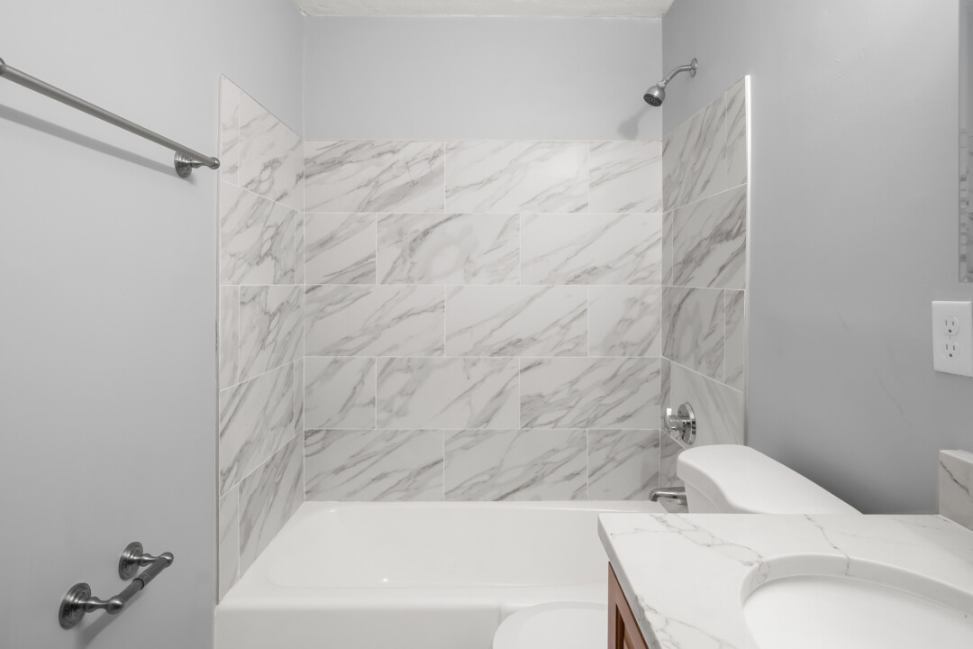 Bright bathroom with a bathtub and shower, toilet, vanity, and marble-patterned walls.