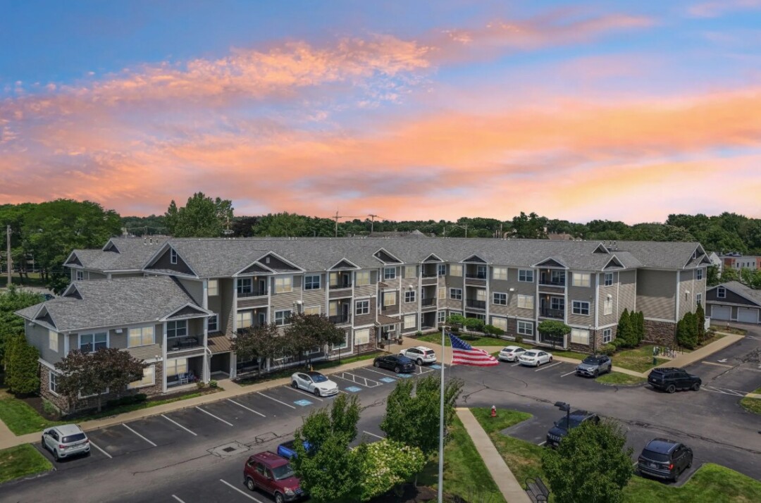 Exterior view of a multi-story residential community with landscaped grounds under a colorful sunset sky.