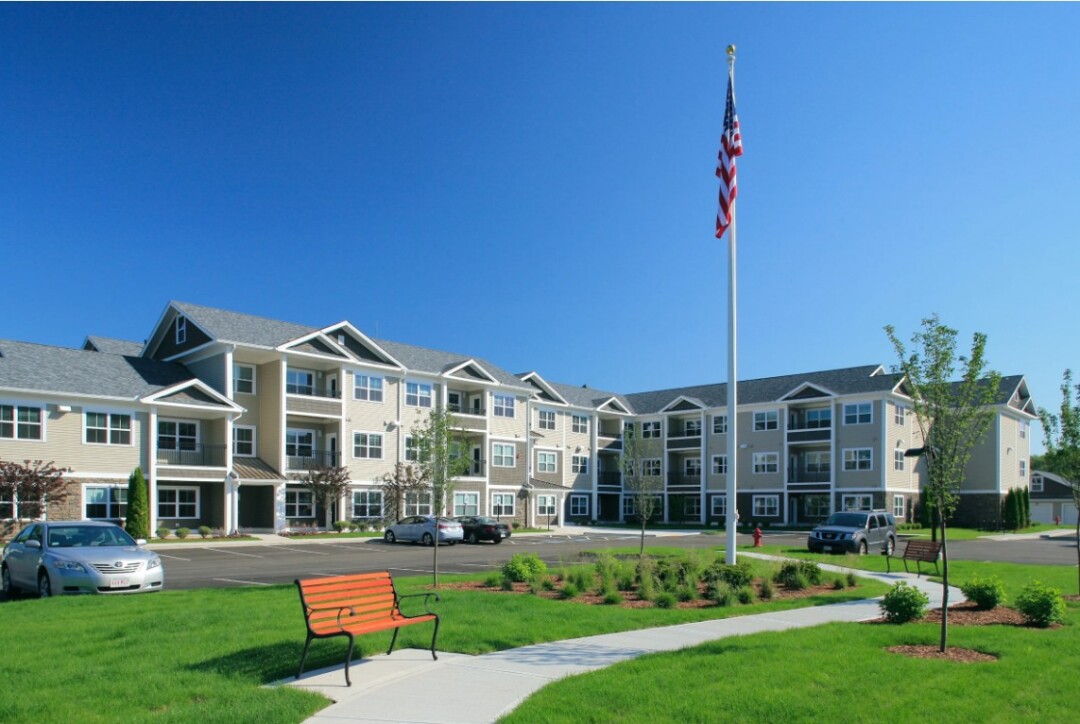 Exterior view of a residential building with a landscaped lawn, walking path, bench, and American flag.