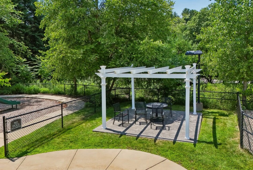 Outdoor seating area with a white pergola, table and chairs, surrounded by trees and greenery.