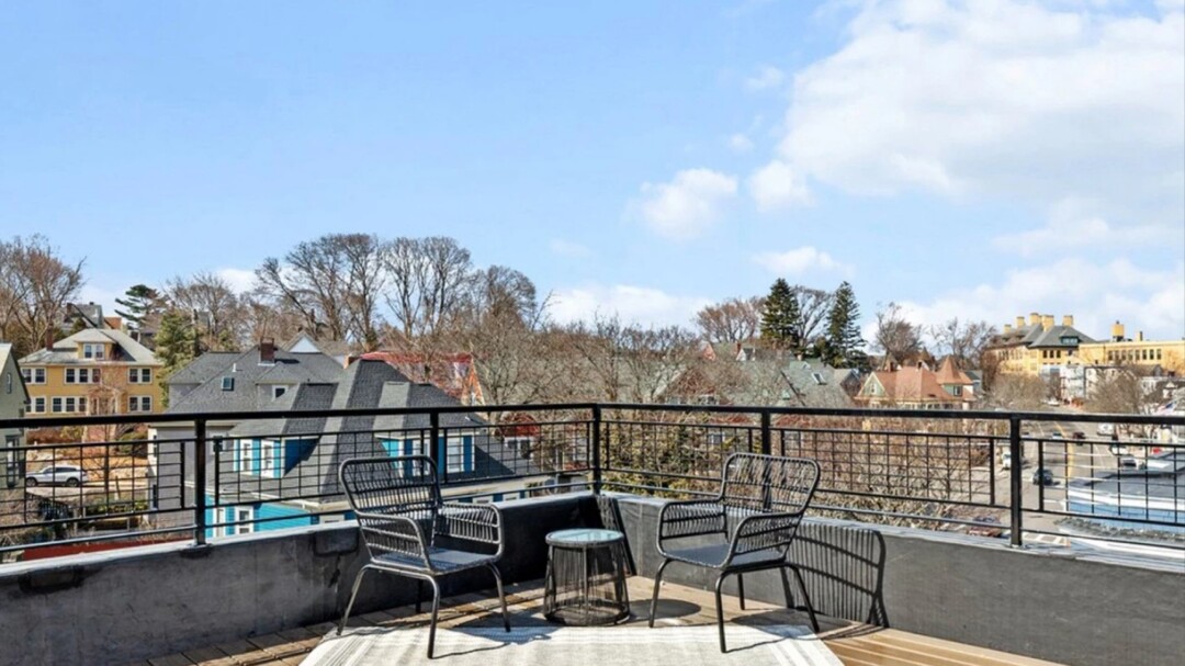 Rooftop terrace with outdoor chairs and a small table overlooking nearby homes, trees, and the surrounding neighborhood.