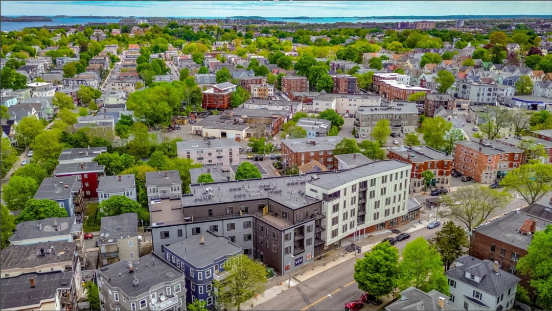 Aerial view of a modern multi-story residential building surrounded by homes, trees, streets, and the surrounding neighborhood.