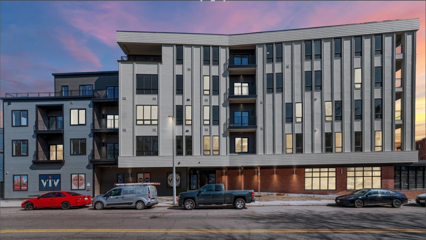Front exterior of a modern multi-story residential building with gray siding, large windows, balconies, and street parking at sunset.