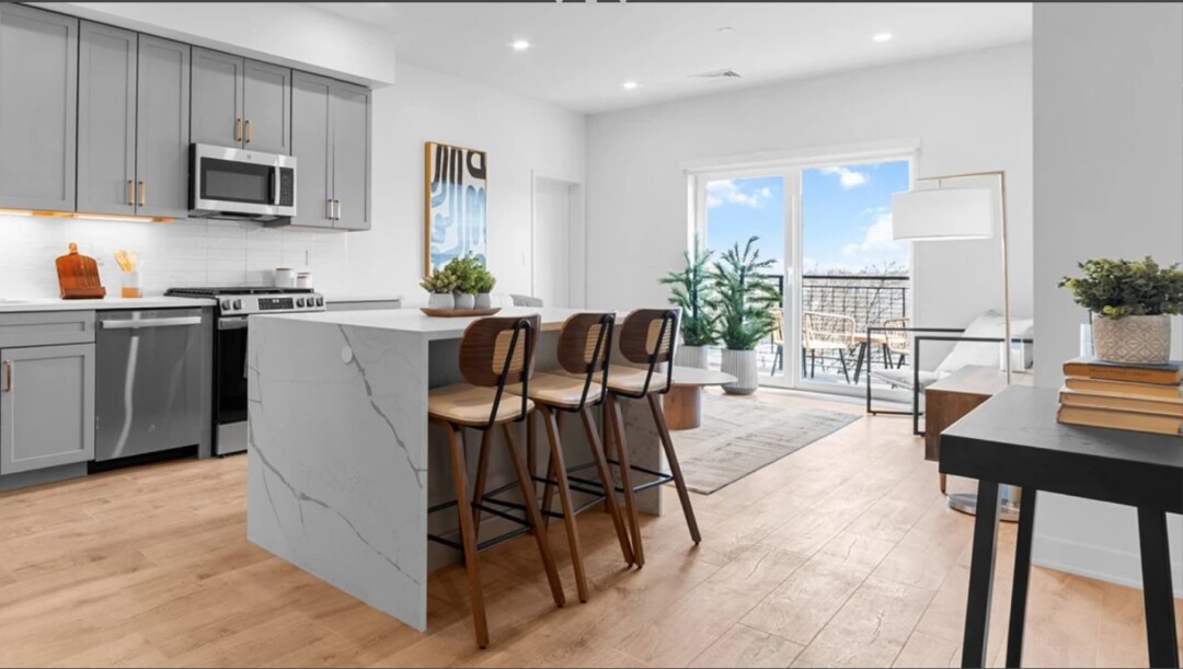 Open-concept kitchen and living room with gray cabinets, stainless steel appliances, a large white island with stools, light wood flooring, and glass doors leading to a balcony.