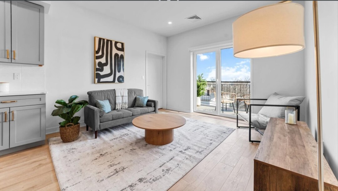 Bright living room with a gray sofa, accent chair, round wood coffee table, area rug, light wood flooring, and glass doors leading to a balcony.