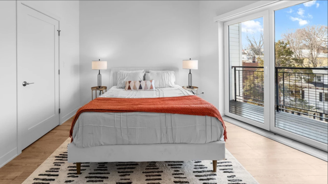 Bright bedroom with a bed, two bedside tables with lamps, light wood flooring, and large glass doors overlooking a balcony.