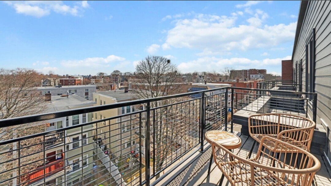 Private balcony with two wicker chairs and a small table overlooking nearby homes and trees under a blue sky.