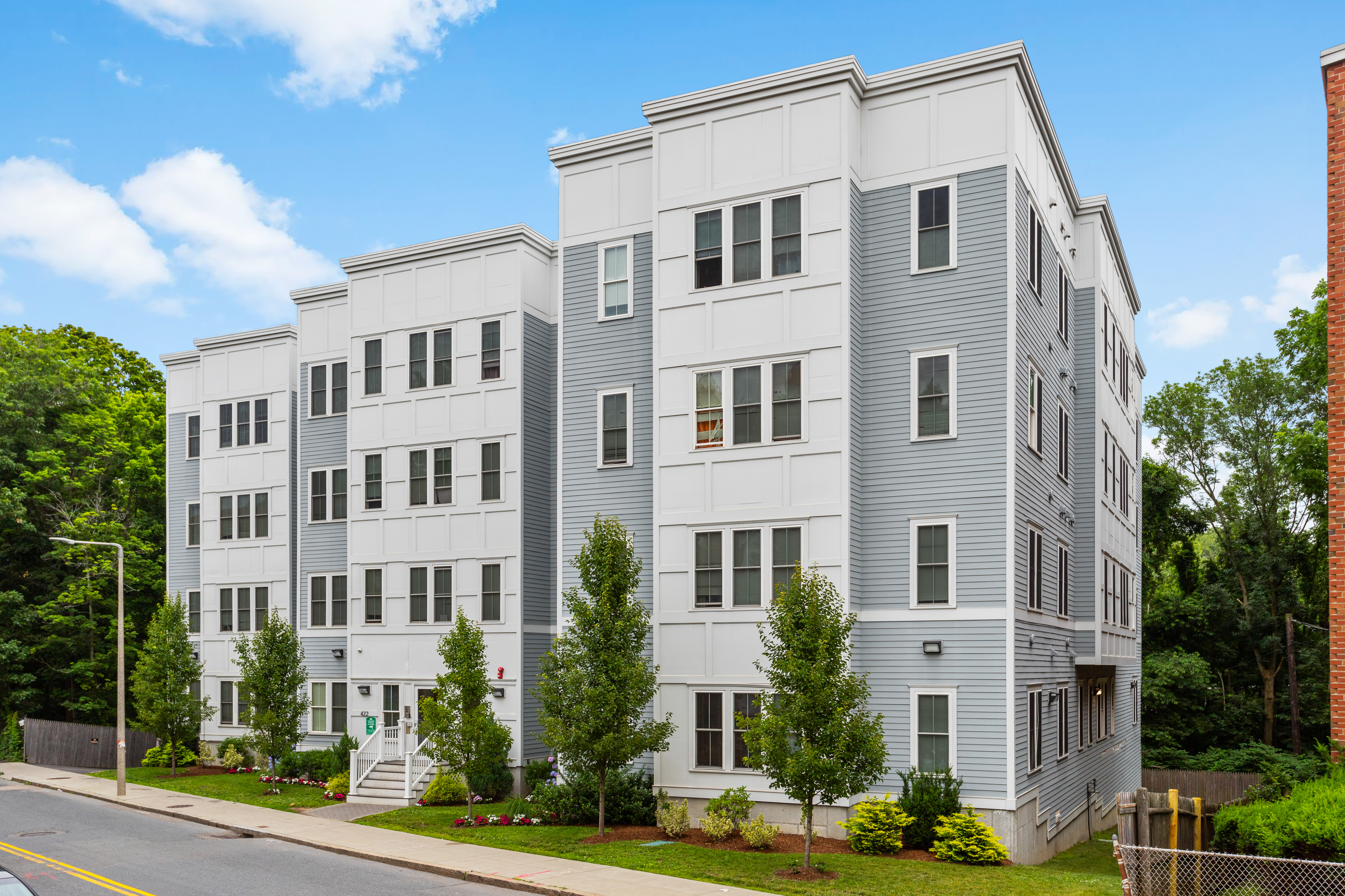 Corner view of a multistory apartment building with white and gray siding, rows of windows, and landscaped shrubs along the sidewalk.