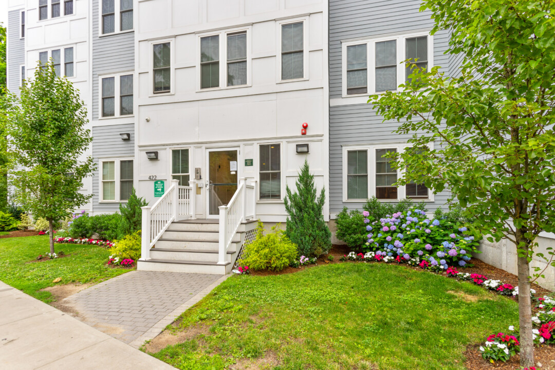 Wider view of the apartment building’s front entrance, showing the steps, white railings, lawn, and landscaped flower beds.