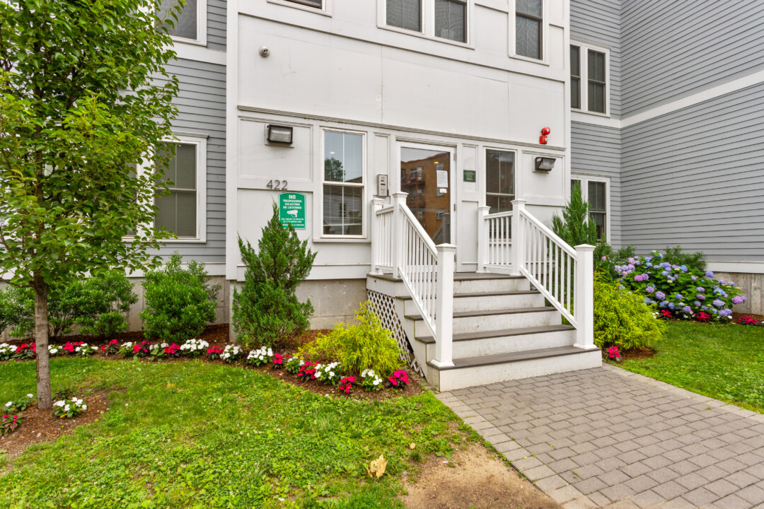Front entrance of a white and gray apartment building, with steps, white railings, a paved walkway, and flower beds.
