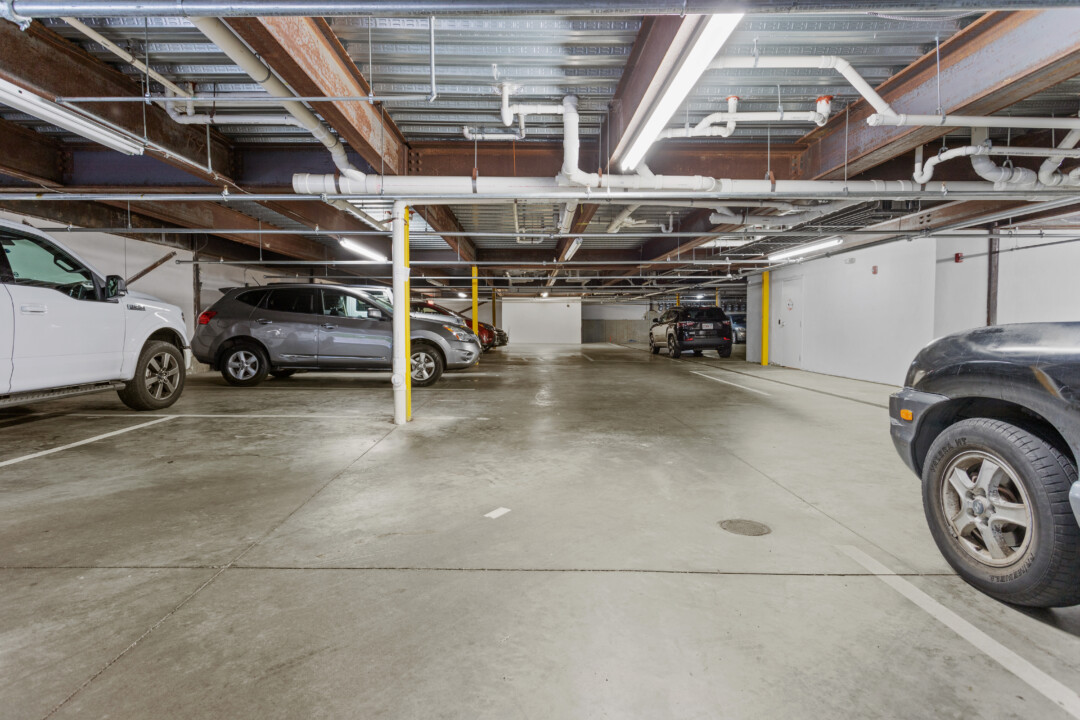 Covered parking garage with concrete flooring, exposed ceiling pipes, support columns, and parked cars.