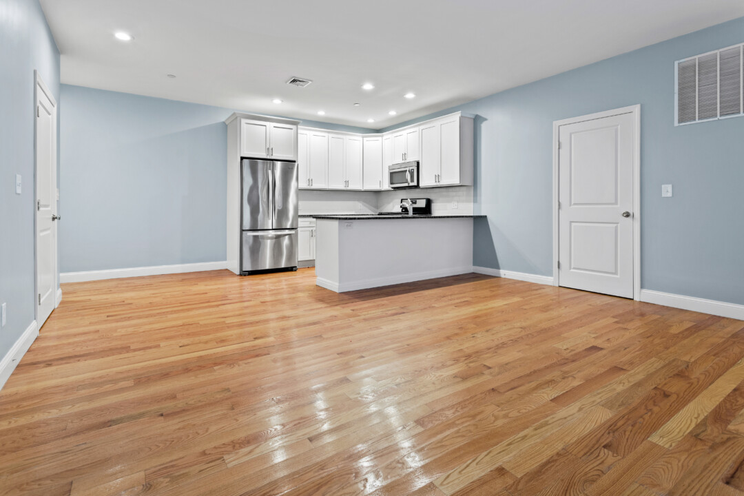 Empty apartment living area with hardwood floors and pale blue walls, opening to a white kitchen with a peninsula and stainless steel refrigerator.
