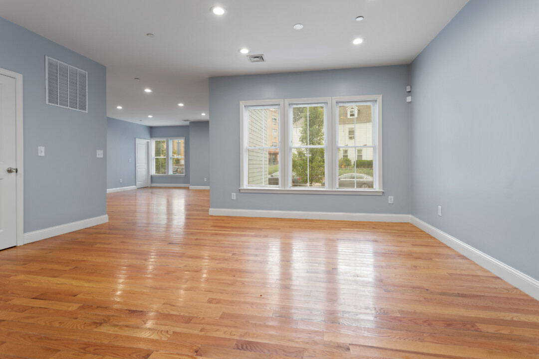 Spacious living area with hardwood floors, pale blue walls, and a large window; another room is visible beyond.